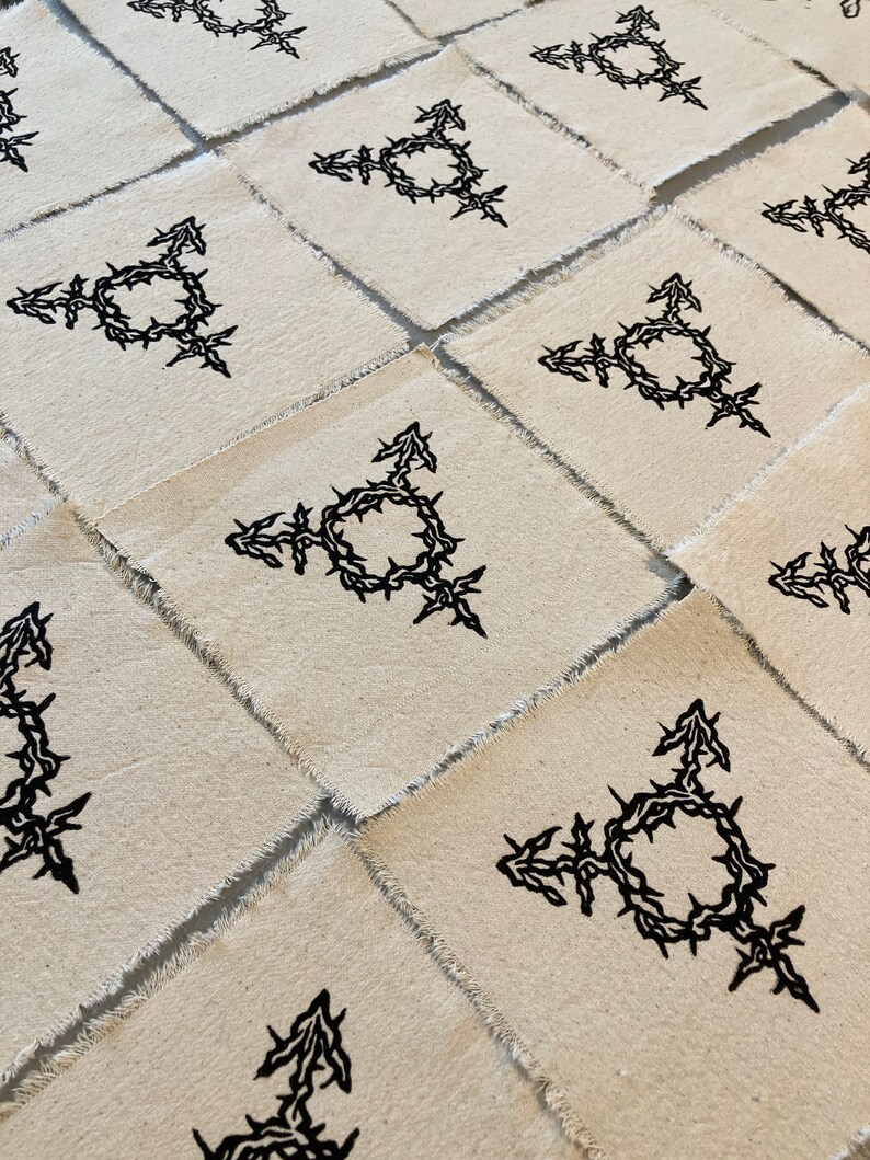 K&ouml;nnte beinhalten: Mehrere quadratische Stoffflicken mit rohen Kanten, die jeweils ein schwarzes Transgender-Symbol zeigen. Das Symbol ist eine Kombination aus m&auml;nnlichen und weiblichen Symbolen und einem Kreis, der mit einem dornigen Rankenmuster verflochten ist.