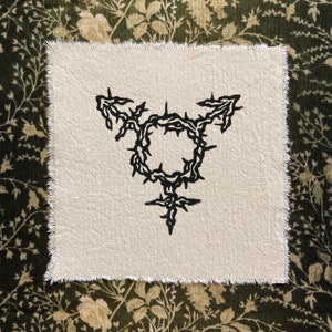 K&ouml;nnte beinhalten: Quadratischer, cremefarbener Stofffleck mit einem schwarzen Transgender-Symbol, das von einem dornigen Kranz umgeben ist. Der Aufn&auml;her hat einen rohen, unfertigen Rand. Der Hintergrund ist ein dunkelgr&uuml;nes und beiges Blumenmuster.