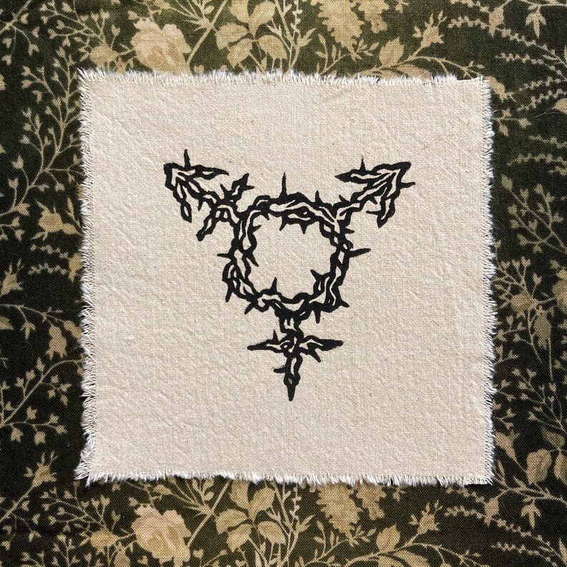 K&ouml;nnte beinhalten: Quadratischer, cremefarbener Stofffleck mit einem schwarzen Transgender-Symbol, das von einem dornigen Kranz umgeben ist. Der Aufn&auml;her hat einen rohen, unfertigen Rand. Der Hintergrund ist ein dunkelgr&uuml;nes und beiges Blumenmuster.