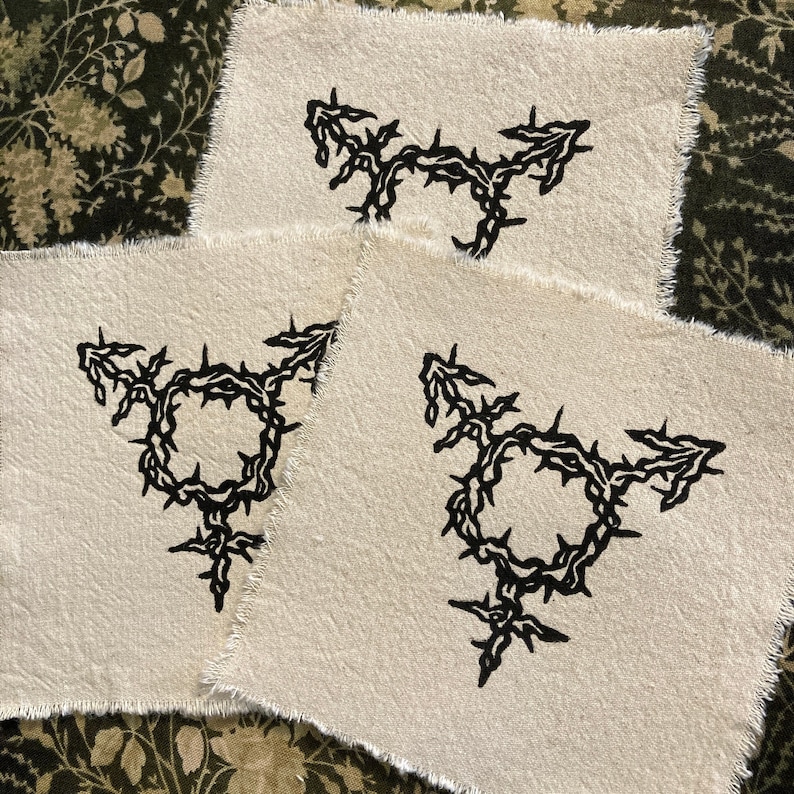 K&ouml;nnte beinhalten: Drei cremefarbene Stoffflicken mit einem schwarzen Transgender-Symbol-Design. Das Symbol ist ein Kreis mit Pfeilen, die nach oben und zu den Seiten zeigen, und einem Kreuz unten, alles umgeben von Dornen.
