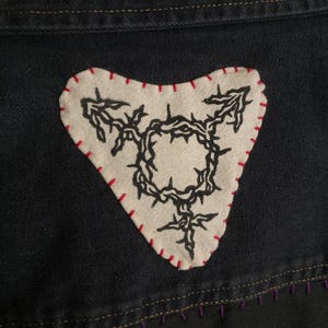 K&ouml;nnte beinhalten: Ein herzf&ouml;rmiger Aufn&auml;her mit einem schwarzen Transgender-Symbol auf cremefarbenem Hintergrund, mit rotem Faden gen&auml;ht. Der Aufn&auml;her befindet sich auf einem dunkelblauen Denim-Stoff.