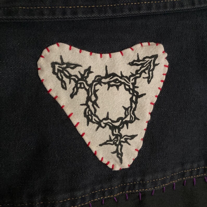 K&ouml;nnte beinhalten: Ein herzf&ouml;rmiger Aufn&auml;her mit einem schwarzen Transgender-Symbol auf cremefarbenem Hintergrund, mit rotem Faden gen&auml;ht. Der Aufn&auml;her befindet sich auf einem dunkelblauen Denim-Stoff.