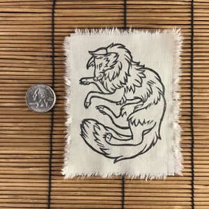 Może przedstawiać: Kwadratowa, kremowa naszywka z czarnym rysunkiem liniowym stylizowanego wilka. Wilk jest zwinięty. Krawędzie naszywki są postrzępione. Obok naszywki znajduje się moneta.