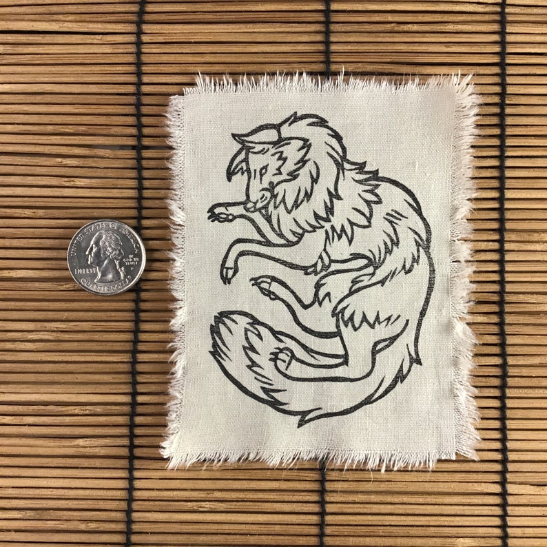 Może przedstawiać: Kwadratowa, kremowa naszywka z czarnym rysunkiem liniowym stylizowanego wilka. Wilk jest zwinięty. Krawędzie naszywki są postrzępione. Obok naszywki znajduje się moneta.