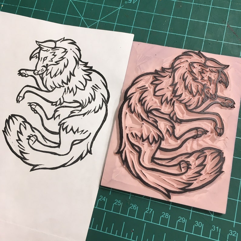 Może przedstawiać: Czarno-biały linoryt stylizowanego wilka leżącego obok r&oacute;żowego bloku linoleum użytego do stworzenia odbitki. Wilk ma szczeg&oacute;łowe futro i jest w zrelaksowanej pozie. Odbitka jest na białym papierze.