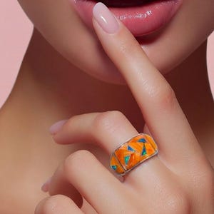 May include: A wide silver band ring featuring a mosaic inlay of vibrant orange and bright turquoise triangular segments. The ring has a polished metallic finish and a geometric pattern that wraps around the entire circumference of the band.