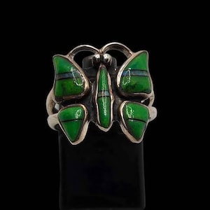May include: A silver ring featuring a butterfly design with inlaid green stone segments. The wings and central body are accented with thin horizontal dark lines and small blue stone slivers, set within a polished metal frame against a dark background.