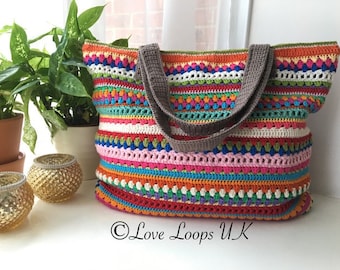 May include: A colorful striped crochet tote bag with brown handles. The bag is made with a variety of stitches and colors, including red, orange, yellow, green, blue, purple, and white. The bag is sitting on a white surface with two gold candle holders.