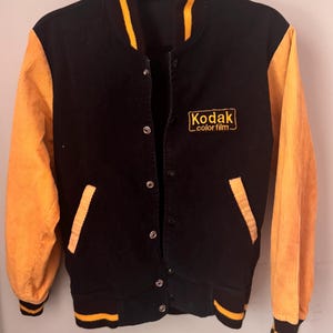 Puede incluir: Una chaqueta universitaria de pana negra con mangas de pana amarilla. La chaqueta presenta un logotipo bordado en amarillo de Kodak color film en el pecho izquierdo, cierre frontal con botones a presión y ribetes acanalados a rayas amarillas y negras.