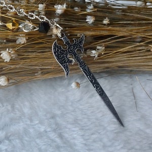 May include: A silver chain necklace with black beads and a pendant shaped like an axe with a detailed design. The pendant is hanging against a background of dried flowers.