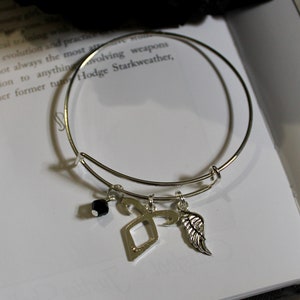 May include: Silver charm bracelet with a black bead, a silver angel wing charm, and a silver rune charm. The bracelet is lying on a white book with text.