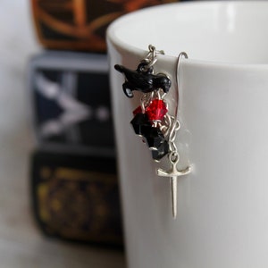 May include: Silver dangle earrings with a black crow charm, black and red beads, and a silver sword charm.