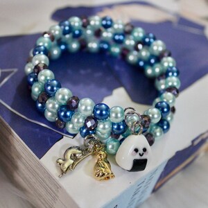 May include: A blue and green beaded bracelet with three charms: a dolphin, a key, and a cartoon onigiri. The bracelet is on a book with a blue cover.