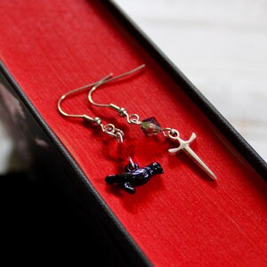 May include: A pair of silver earrings with a black raven charm and a silver sword charm. The earrings are hanging from a red book.