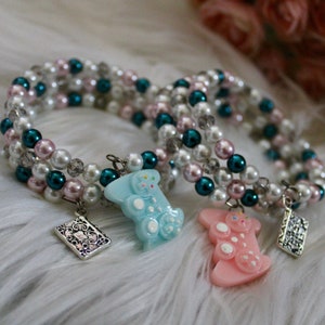 May include: Three beaded bracelets with charms. The bracelets are made with white, pink, and teal beads. The charms are blue and pink gummy bear shapes.