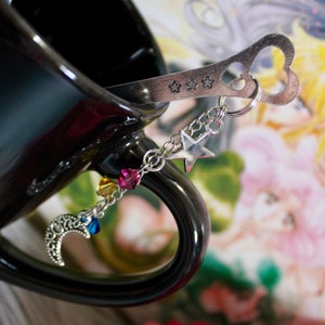 May include: A black ceramic mug with a silver spoon attached to a charm bracelet. The bracelet features a crescent moon, stars, and colorful crystals.