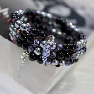 May include: A multi-strand bracelet made with black, silver, and purple beads. The bracelet has two silver charms in the shape of wings.