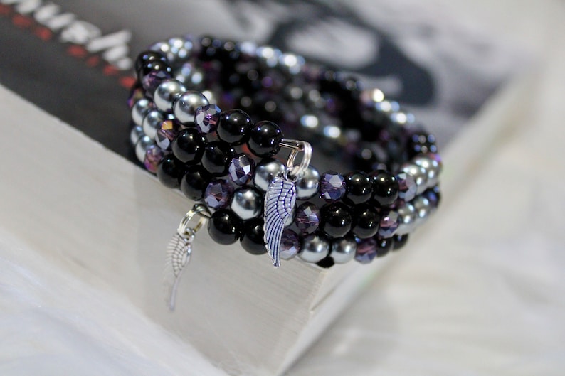 May include: A multi-strand bracelet made with black, silver, and purple beads. The bracelet has two silver charms in the shape of wings.