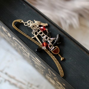 May include: A silver bookmark with a chain and charms, including a gun, a sword, a raven, a wine glass, and red and black crystals.