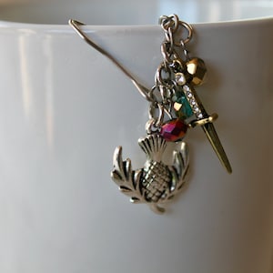 May include: A silver charm bracelet with a thistle charm, a sword charm, a gold bead, and a green crystal charm. The bracelet is hanging from a silver hair clip.