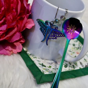 May include: A iridescent butterfly-shaped spoon charm hangs from a white ceramic mug with a green and white floral coaster. The spoon has a flower design on the bowl and is iridescent.