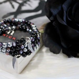 May include: A multi-strand bracelet with black, silver, and purple beads. The bracelet has two silver angel wing charms.