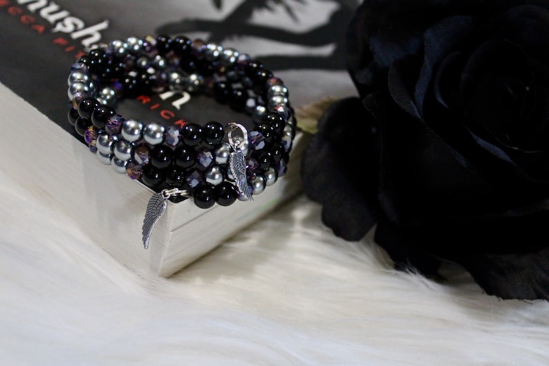 May include: A multi-strand bracelet with black, silver, and purple beads. The bracelet has a silver charm in the shape of a wing.