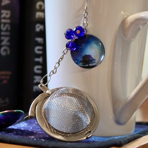 May include: A silver tea infuser with a mesh ball and a chain attached to a blue glass charm with a nature scene. The charm has a silver rim and is decorated with blue crystal beads.