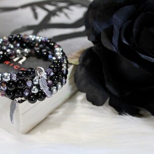 May include: A multi-strand bracelet with black, silver, and purple beads. The bracelet has two silver angel wing charms.