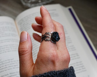 May include: A silver ring with a black gemstone setting. The ring is on a person's finger and the hand is resting on a book.