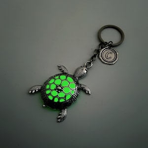 May include: A silver turtle keyring with a glow-in-the-dark green shell. The keyring has a silver ring with a silver charm that says "C".