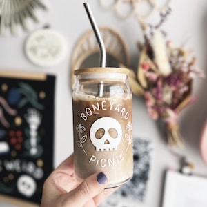 Peut inclure: Un verre à café transparent avec un couvercle en bambou et une paille en acier inoxydable. Le verre porte les mots "BONEYARD PICNIC" et un motif de crâne blanc. Le verre est rempli d'une boisson brun clair.