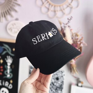 May include: Black baseball cap with white embroidery that reads "SERIOUS" with a small white daisy with a yellow center.
