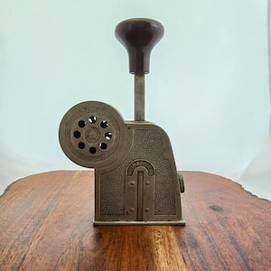 May include: A vintage metal number stamp with a black plastic handle. The stamp has a round dial with numbers and a lever to adjust the number. The stamp is sitting on a wooden surface.