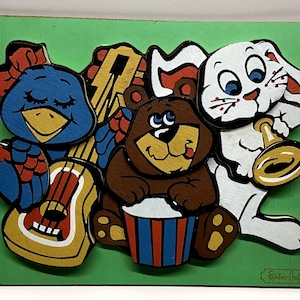 Può includere: Un puzzle in legno per bambini con un uccello blu, un orso marrone e un coniglio bianco che suonano strumenti musicali. L'uccello ha un fiocco rosso, l'orso suona un tamburo e il coniglio suona una tromba. Lo sfondo è verde.