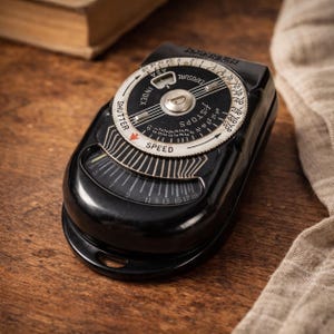Könnte beinhalten: Ein schwarzer Ranger Belichtungsmesser im Vintage-Stil mit weißen und silbernen Zifferblättern. Das Messgerät hat ein kreisförmiges Zifferblatt mit Blenden- und ASA-Einstellungen und eine Nadel, die auf die Geschwindigkeit zeigt. Auf einer Holzoberfläche.