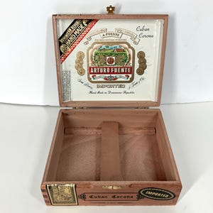 May include: An open wooden cigar box with a hinged lid. The lid features a printed label with the text "ARTURO FUENTE" and "IMPORTED". The box is empty, showing the interior construction. The box is labeled "Cuban Corona" and "Hand Made".
