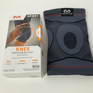 Könnte beinhalten: Eine graue McDavid Knie-Kompressionsstrickhülse mit Gelpolster und eine passende Box. Die Box zeigt das McDavid-Logo, das Wort "SPORT" und den Text "KNEE COMPRESSION KNIT SLEEVE w/ gel buttress". Die Hülse ist für moderate Unterstützung und Schmerzlinderung im Knie konzipiert.
