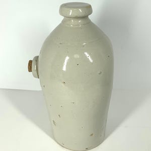 Puede incluir: Una jarra de gres de color gris claro. Tiene un cuerpo redondeado, un cuello estrecho y un pequeño tapón redondo. Un corcho está insertado en el lateral. Es probable que sea una pieza antigua.