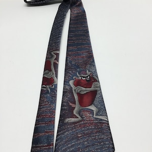 May include: A blue silk tie with a red and white cartoon Tasmanian Devil print. The devil is standing with its arms crossed and has a red heart on its chest.