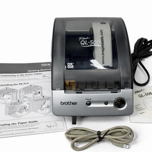 Puede incluir: Impresora de etiquetas Brother QL-500 en negro y plateado con un rollo de etiquetas visible. Tiene un botón de encendido y un botón de avance. Incluye un cable de alimentación, un CD y un manual de usuario.