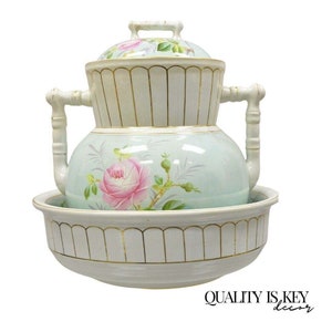 May include: A three-tiered porcelain water pitcher with a floral design. The pitcher has a light blue background with pink roses and green leaves. The top tier has a lid with a gold trim. The bottom tier is a bowl with a gold trim.