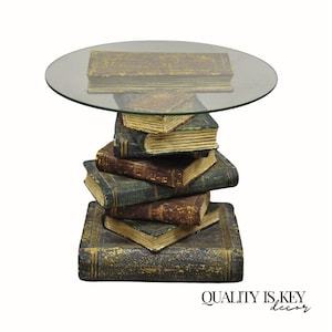 May include: A unique side table with a clear glass top supported by a stack of faux antique books. The books are in shades of brown, green, and gold, creating a vintage aesthetic. The table is designed for decorative use.