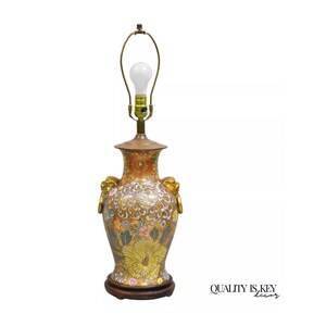 May include: A gold and orange ceramic table lamp with floral designs and gold accents. The lamp has a round base and two gold handles on the sides. The lampshade is missing.