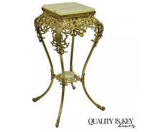 May include: A gold metal plant stand with an ornate design and a white marble top. The stand has three legs and a round base.