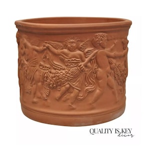 May include: A terracotta-colored planter with a detailed relief of figures in a classical style. The cylindrical pot features a wide rim and a decorative band of sculpted cherubs and other figures around its exterior. The words "QUALITY IS KEY decor" are at the bottom.