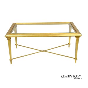 May include: A rectangular gold coffee table with a glass top. The table has four legs with an X-shaped support and decorative detailing. The frame is a bright gold color. The table is a classic design.