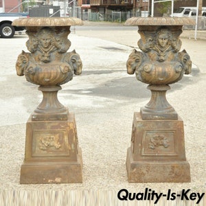 May include: Two antique cast iron urns with detailed relief carvings. Each urn features a female face and ram heads, set atop a square pedestal base. The urns have a weathered, aged appearance, suggesting outdoor use. The overall color is a muted bronze.
