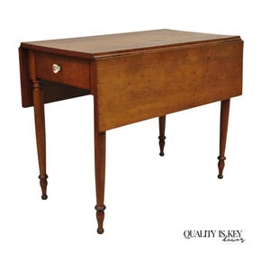 May include: A vintage wooden drop-leaf table with a warm brown finish. The table features a single drawer with a crystal knob and slender, turned legs. The table's design is classic and elegant, suitable for various interior styles.