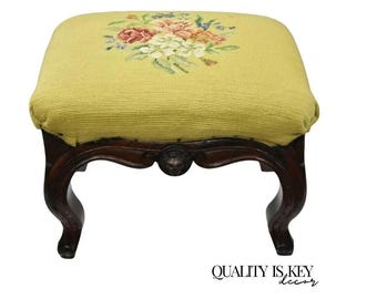 May include: An antique footstool with a light yellow upholstered top, decorated with a floral needlepoint design in pink, white, and green. The dark wood frame has ornate, curved legs and a decorative apron. The text "QUALITY IS KEY decor" is at the bottom.